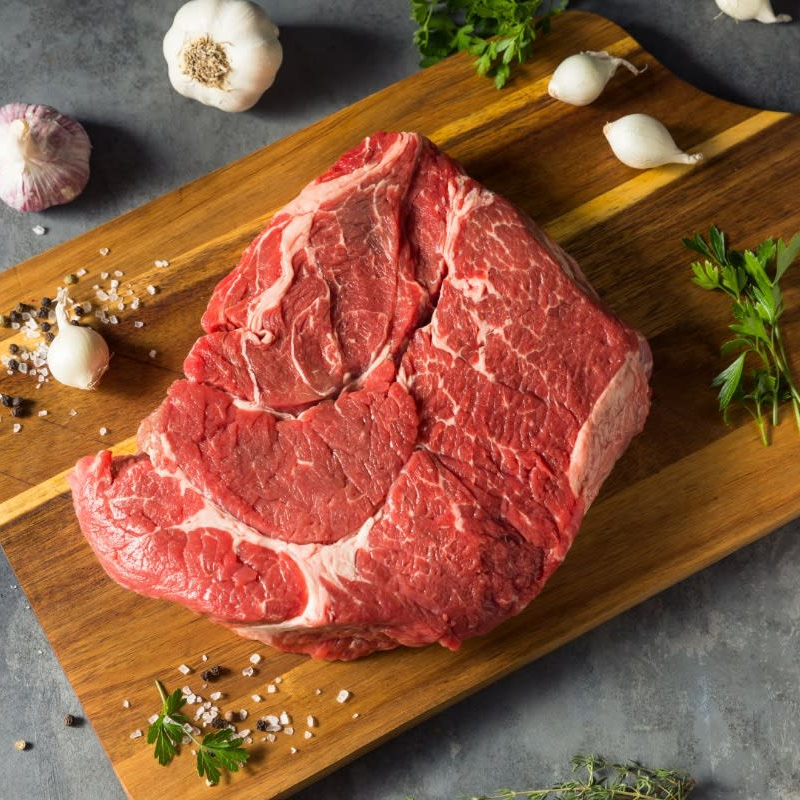 Raw chuck roast steak on a wooden cutting board with herbs and garlic on a gray surface