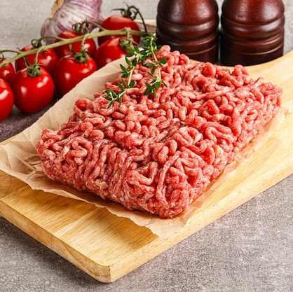 Raw ground meat on a wooden cutting board with tomatoes and pepper mills in the background.